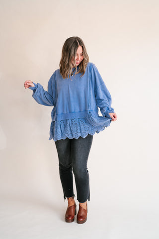A woman modeling a dusty blue long-sleeve top with a scalloped eyelet lace hem, paired with black jeans and brown leather heels, against a plain off-white studio background.