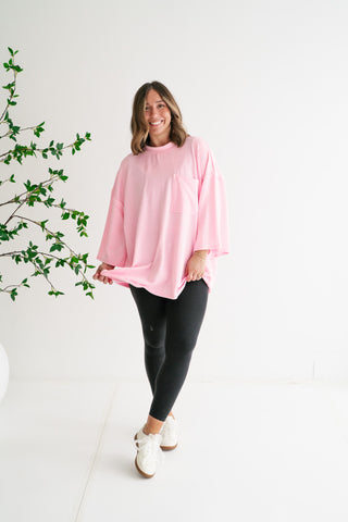 A woman smiling in a bright studio, wearing an oversized blush pink ribbed tunic sweater with a front pocket, paired with black leggings and white sneakers.