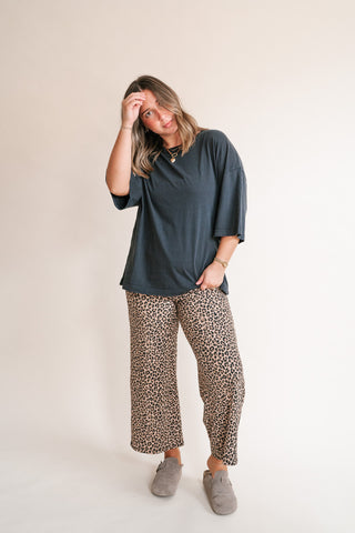 A woman modeling a charcoal short-sleeve top and leopard-print wide-leg pants with gray slip-on shoes, standing against a plain off-white studio background.