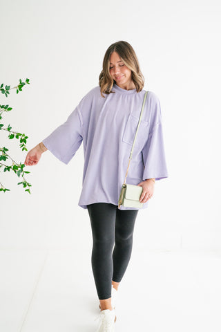 A woman modeling an oversized lavender ribbed-knit tunic with wide sleeves and a front pocket, paired with black leggings and white sneakers in a bright studio.