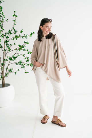 A woman smiling in a bright studio wearing an oversized tan ribbed waffle-knit tunic, white wide-leg jeans, and tan sandals.