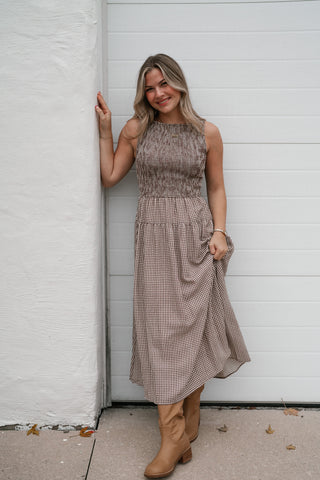 Model leaning against a wall in the brown gingham sleeveless midi dress and tan western boots