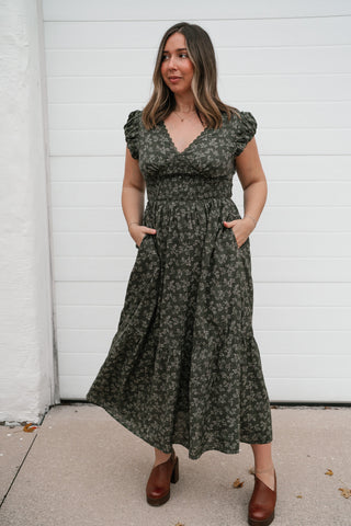Model with hands in the pockets of the olive floral V-neck midi dress