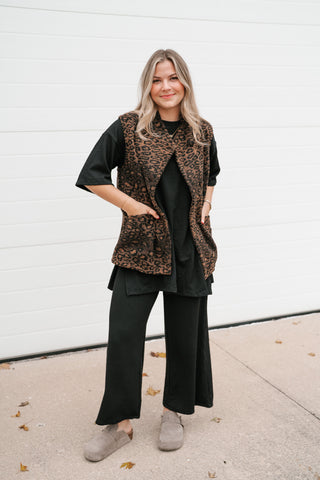 Full-length front view of the relaxed leopard print puffer vest worn open over black wide-leg pants and suede booties