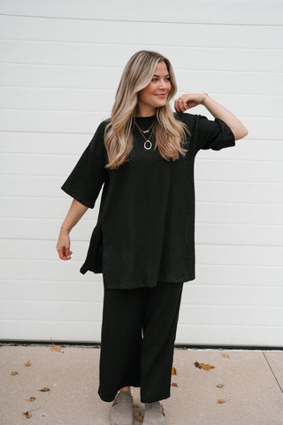 Smiling model in the black ribbed oversized top showing the dolman sleeves and side slits