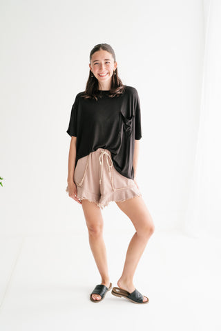 A woman smiling in a bright studio wearing an oversized black pocket t-shirt and dusty rose ruffled lounge shorts with a drawstring waist.
