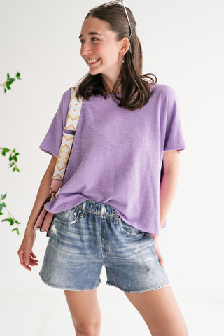 A woman smiling and wearing a lavender-colored textured oversized t-shirt paired with light-wash denim shorts and a pink crossbody bag.