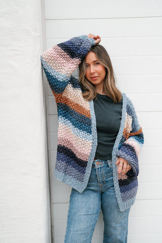 Woman leaning against a white wall wearing the Something Special oversized striped chunky crochet open cardigan in slate blue, blush, rust and navy with jeans