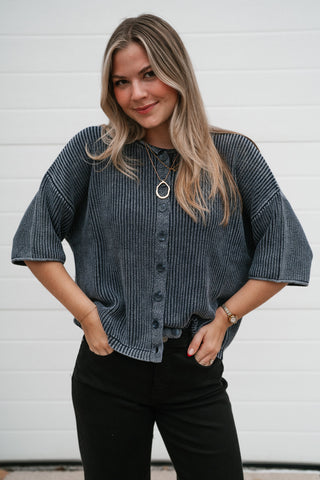 Model with hands in pockets wearing the boxy navy ribbed cardigan buttoned up with black pants