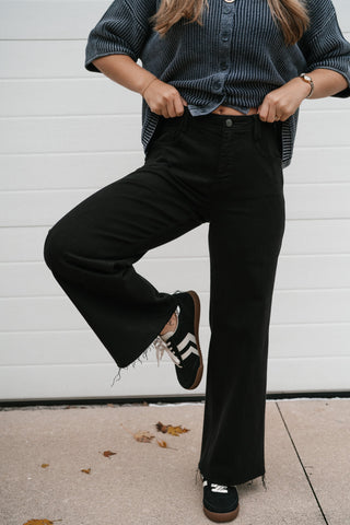 Model lifting one knee to show the stretch and wide leg of the black Risen high rise ankle jeans