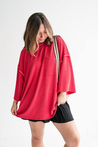 A woman modeling an oversized, vibrant red ribbed mock-neck top featuring exposed white stitching and a front pocket, paired with black shorts.