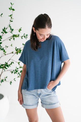 A woman modeling a relaxed, boxy-fit slub cotton t-shirt in dusty blue, paired with light-wash denim shorts in a bright, minimalist studio setting.