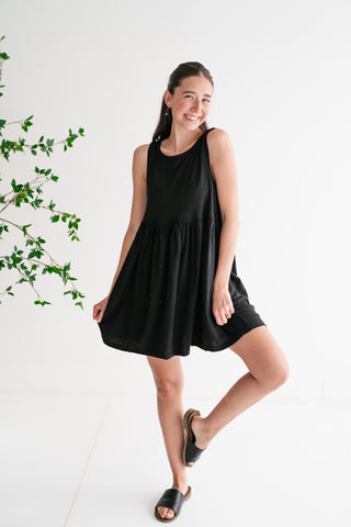 A smiling woman wearing a sleeveless black babydoll mini dress and black leather slides, posing against a clean white background.
