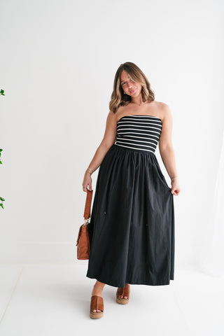 A woman posing in a chic black and white striped strapless maxi dress with a flowy black skirt, carrying a tan leather handbag and wearing platform sandals.