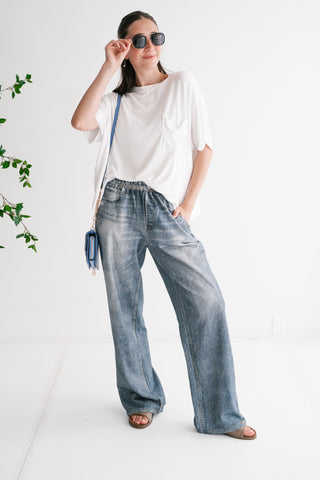 A woman modeling high-waisted, light-wash wide-leg printed denim jeans paired with an oversized white pocket t-shirt and sunglasses in a bright minimalist studio.