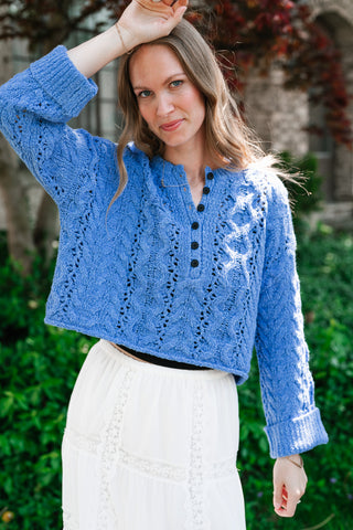 A woman outdoors wearing a vibrant cornflower blue cropped pointelle knit cardigan with dark button-front detailing and a white lace skirt.