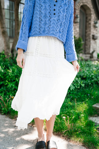 A woman wearing a flowy, white tiered maxi skirt with delicate lace trim, paired with a vibrant blue cable-knit sweater in a lush garden setting.