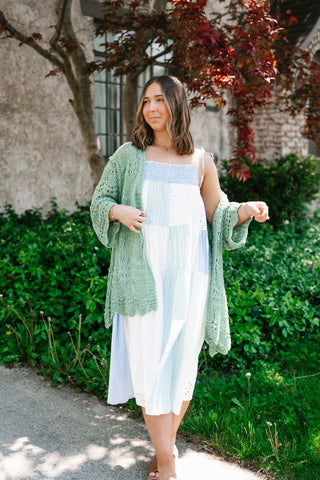 A woman standing outdoors wearing a light blue and white patchwork sundress layered with an oversized, sage green crochet-knit cardigan with intricate lace detailing.