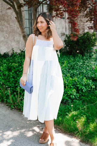 Woman wearing a light blue and white striped tiered midi dress with eyelet embroidery and tie-shoulder straps in a sunny outdoor setting.