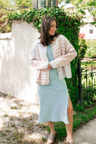 Woman in a light blue dress and striped cardigan standing outdoors next to a white wall with greenery.