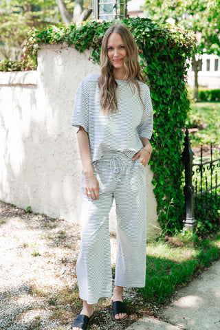 A woman stands outdoors wearing a matching two-piece grey and white horizontal striped lounge set, featuring a boxy short-sleeve top and wide-leg drawstring pants, paired with black slide sandals.