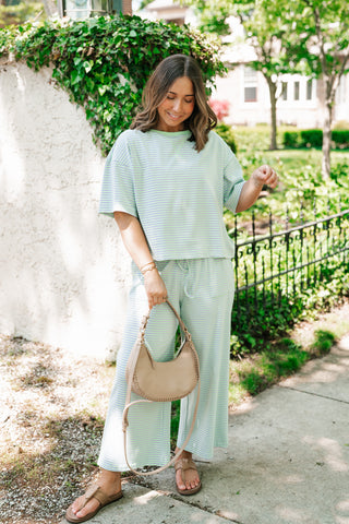A woman smiling outdoors wearing a sage green and white horizontal striped two-piece lounge set featuring a relaxed-fit short-sleeve top and matching wide-leg pants, accessorized with a beige crescent-shaped handbag and tan sandals.
