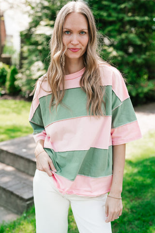 A woman wearing an oversized color-block t-shirt with wide horizontal stripes in pastel pink and sage green, featuring trendy raw-edge exposed seams.