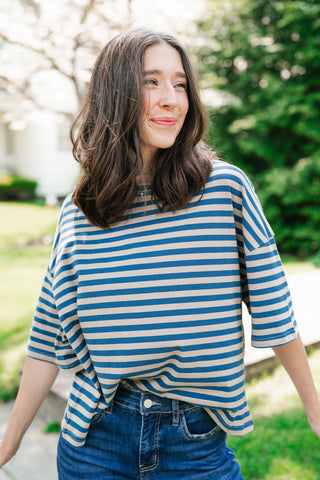 A woman with wavy brown hair wearing an oversized, blue and tan horizontal striped t-shirt paired with denim jeans, smiling in an outdoor setting.