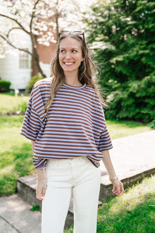 Woman wearing an oversized brown and lavender horizontal striped t-shirt with white jeans in a sunny outdoor garden setting.