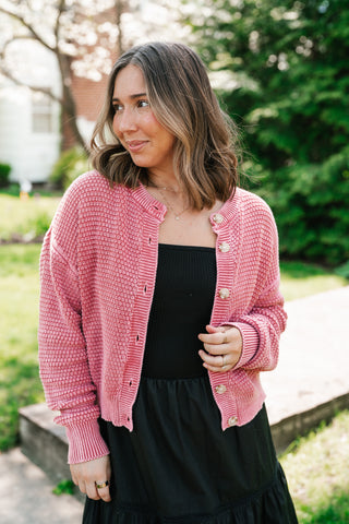 A woman smiling in a garden wearing a dusty rose chunky knit cardigan with cream-colored buttons over a black midi dress.