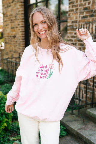 A smiling woman wearing a light pink crewneck sweatshirt featuring a pink tulip and the embroidered text, "What a friend we have in Jesus," standing in front of a brick building.