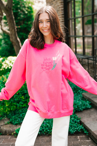 A smiling young woman wearing a vibrant pink oversized sweatshirt featuring the embroidered phrase "What a friend we have in Jesus" alongside a delicate tulip illustration.