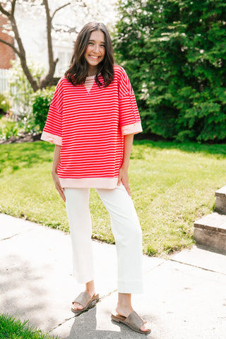 A woman smiling outdoors wearing a vibrant red and pink horizontal striped oversized short-sleeve sweatshirt with white cropped trousers and tan slides.