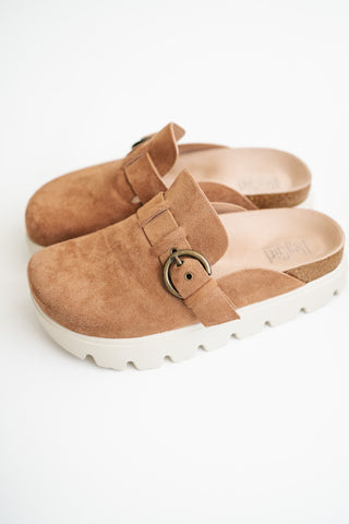 A pair of tan suede clogs with a chunky white sole and a bronze metal ring detail on the strap, shown on a clean white background.