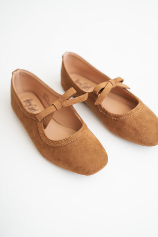 A pair of tan suede Mary Jane flats with bow details on the toes, displayed on a clean white background.