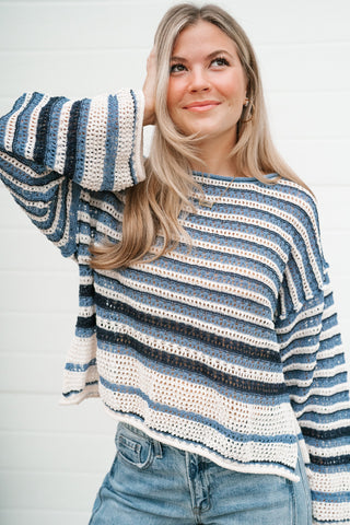 A woman wearing a blue, white, and black striped crochet knit sweater with flared bell sleeves, paired with blue jeans, standing against a white paneled wall.