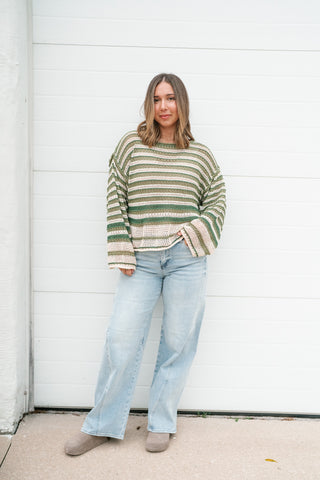 A woman stands in front of a white garage door wearing a green and beige striped knit sweater, light-wash wide-leg jeans, and taupe clogs.