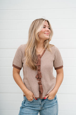 A woman wearing a brown and beige houndstooth short-sleeve top with ruffle trim down the front, paired with blue jeans, standing against a white shiplap wall.