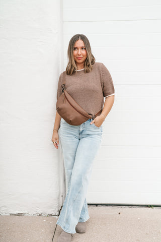 A woman stands against a white paneled wall wearing a brown short-sleeve knit top, light blue jeans, and grey clogs, with a brown crossbody bag slung across her body.