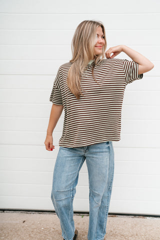 A woman wearing a brown and mint horizontally striped short-sleeve polo shirt and light blue jeans, standing in front of a white garage door.