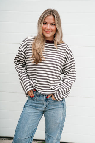 A woman with blonde hair wearing a grey and white striped long-sleeve top and blue jeans, standing in front of a white paneled wall.