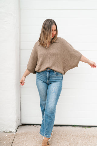 A woman wearing a taupe knit poncho-style top and light-wash blue jeans, standing in front of a white garage door.