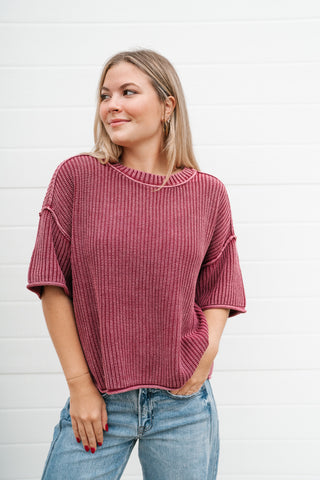 A woman wearing a ribbed knit short-sleeve top in dusty rose pink, paired with light blue jeans, standing against a white shiplap wall.