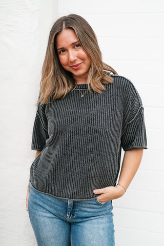 A woman wearing a dark gray ribbed knit short-sleeve top and blue jeans, standing against a white paneled wall.