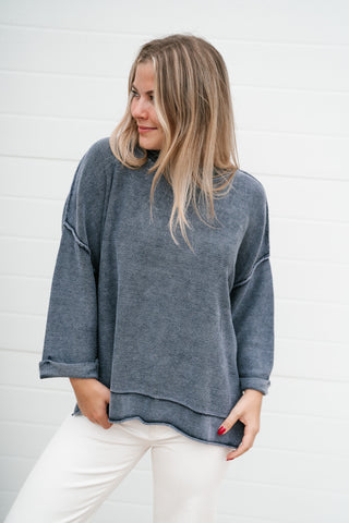A woman wearing a washed slate blue 3/4 length sleeve sweater and white pants, standing against a white paneled wall.