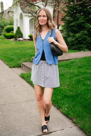 A woman walking on a sidewalk wearing a vibrant azure blue sleeveless knit button-down vest paired with a black and white gingham mini skirt.