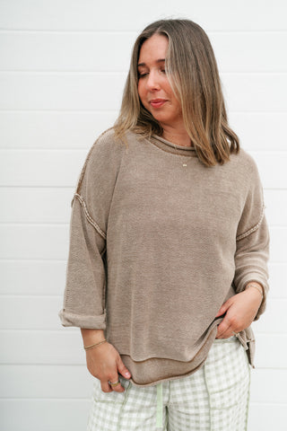 A woman wearing a taupe-brown knit crewneck sweater with rolled sleeves and paired with green and white gingham pants, standing against a white shiplap wall.