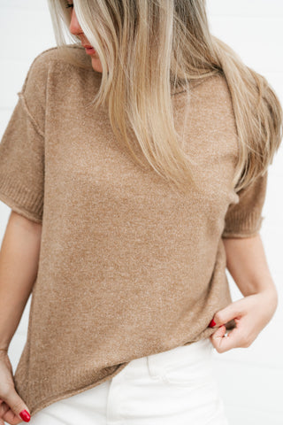 A woman wearing a short-sleeve knit sweater in a warm camel brown color, paired with white pants, shown from a side-back angle against a plain white background.