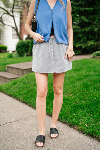 Woman wearing a black and white gingham mini skirt with button-front detail, paired with a blue knit vest and black slides on a sunny sidewalk.