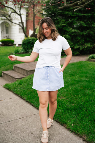 A woman walking outdoors wearing a grey t-shirt, blue and white gingham seersucker button-front mini skirt with pockets, and tan sneakers.